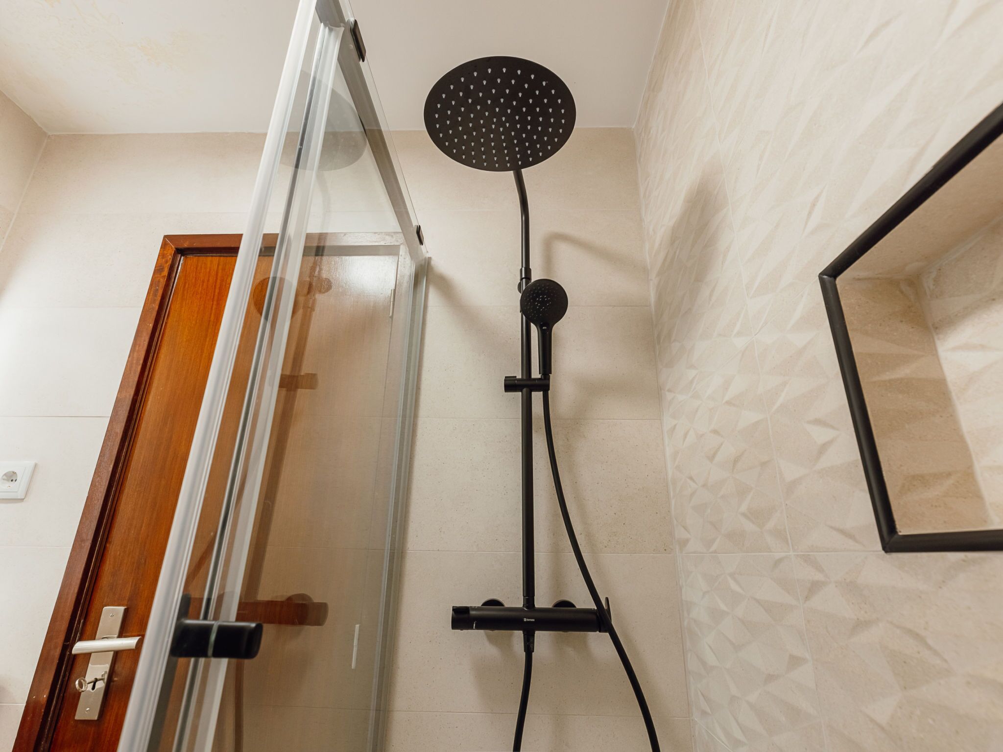 A shower with a handheld showerhead mounted on a wall, a glass door, and a wooden door frame.