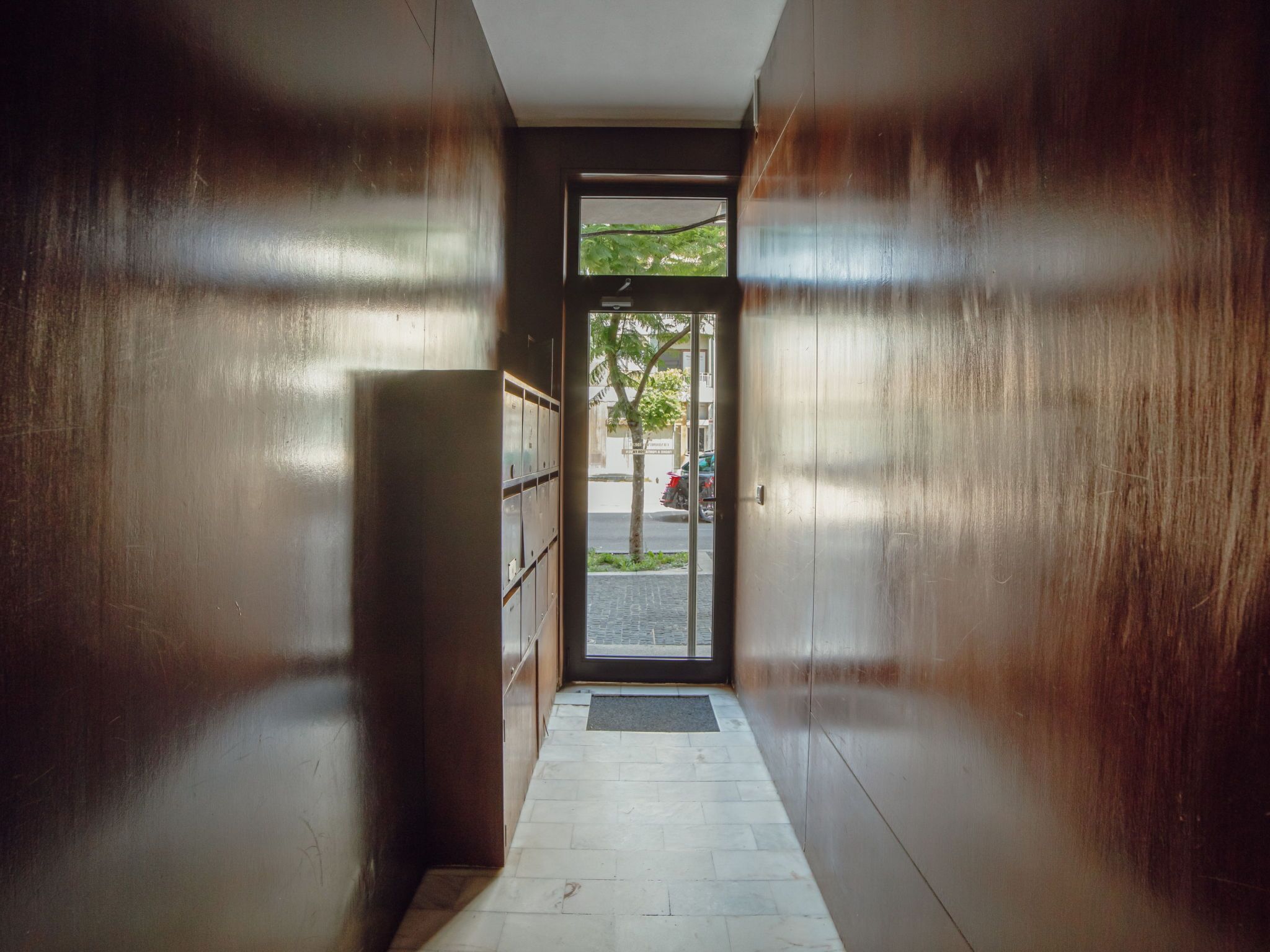 A narrow hallway with a door leading outside, a metal bench on the left, and a tiled floor.