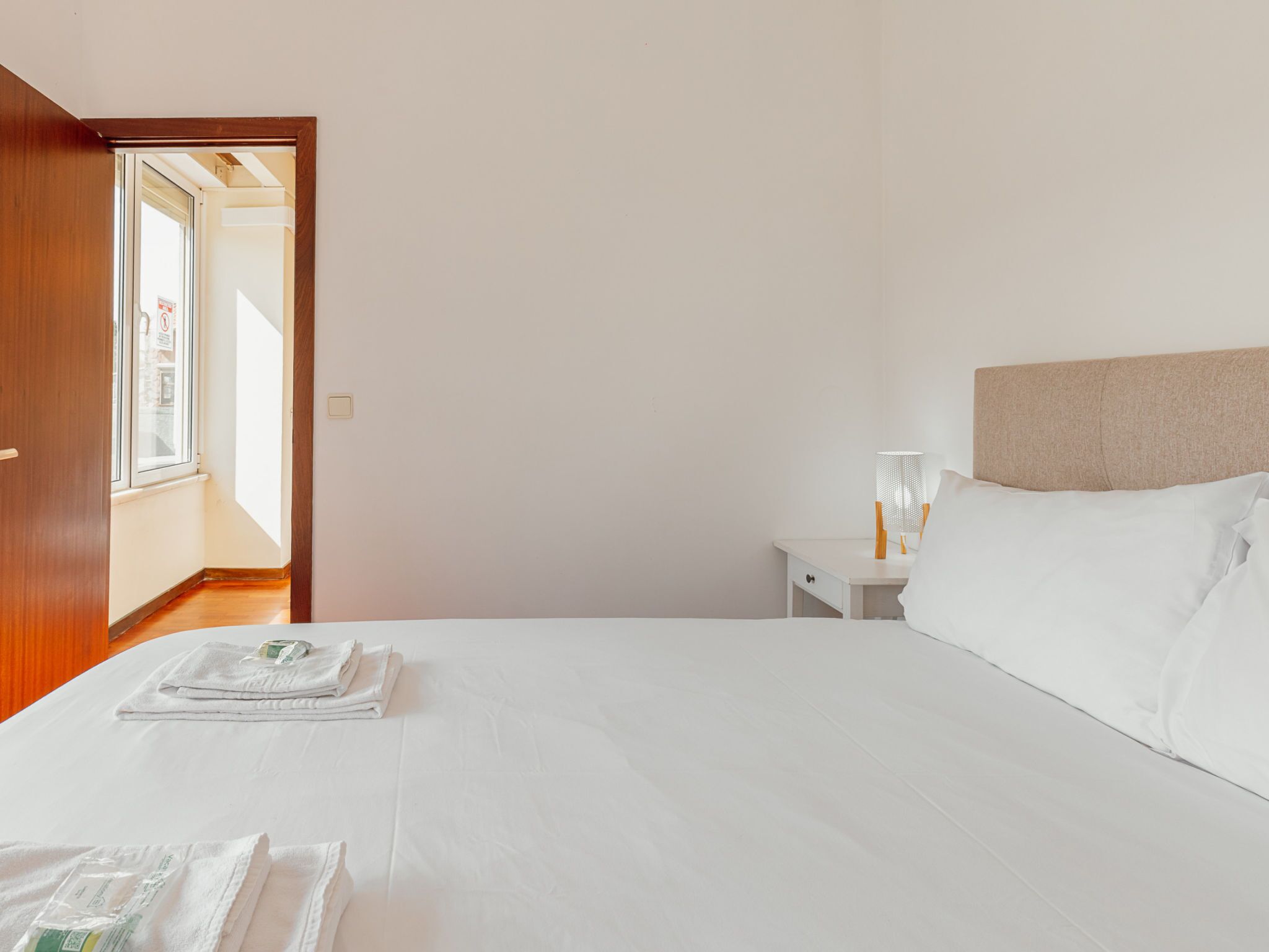 A hotel room with a bed, a nightstand with a lamp, and a door leading to another room.