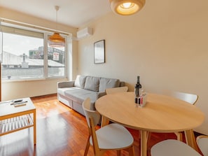 A modern living room with a sofa, a dining table with chairs, a hanging light fixture, and a window with a view of buildings. - Apartment  by Interhome (Porto)