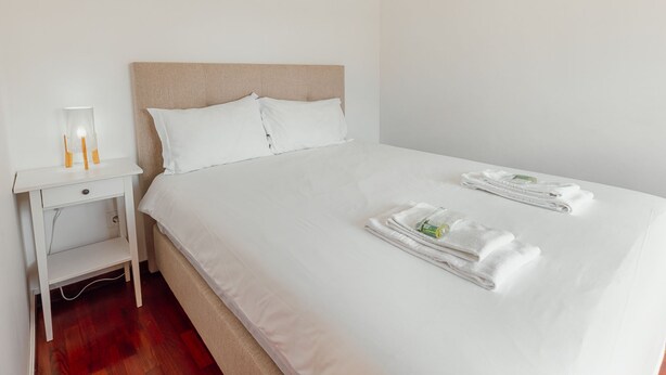 A neatly made bed with white linens and towels, a bedside table with a lamp and a small item on it, and a plain wall. - Apartment  by Interhome (Porto)