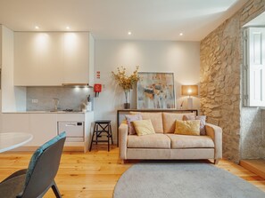 A modern kitchen with white cabinets, a dining area with a round table, a sofa with cushions, and a stone wall. - Apartment  by Interhome (Porto)