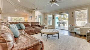 Ein geräumiges Wohnzimmer mit einem braunen Ledersofa, einem runden Hocker, Deckenventilatoren und großen Fenstern.