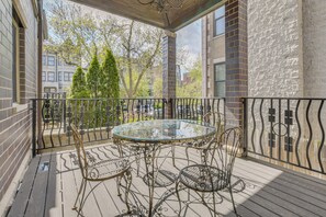 A balcony with a glass table and metal chairs, overlooking a street with trees and buildings.