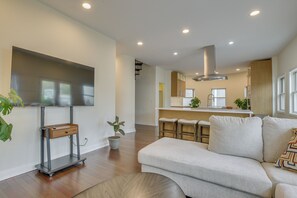 A modern living room with a sofa, a TV on a stand, a kitchen area with a bar, and a staircase.