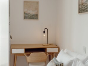 Ein schlicht eingerichteter Raum mit einem Holztisch, einem Stuhl, einem Bett mit weißer Bettwäsche und einer Wandlampe.