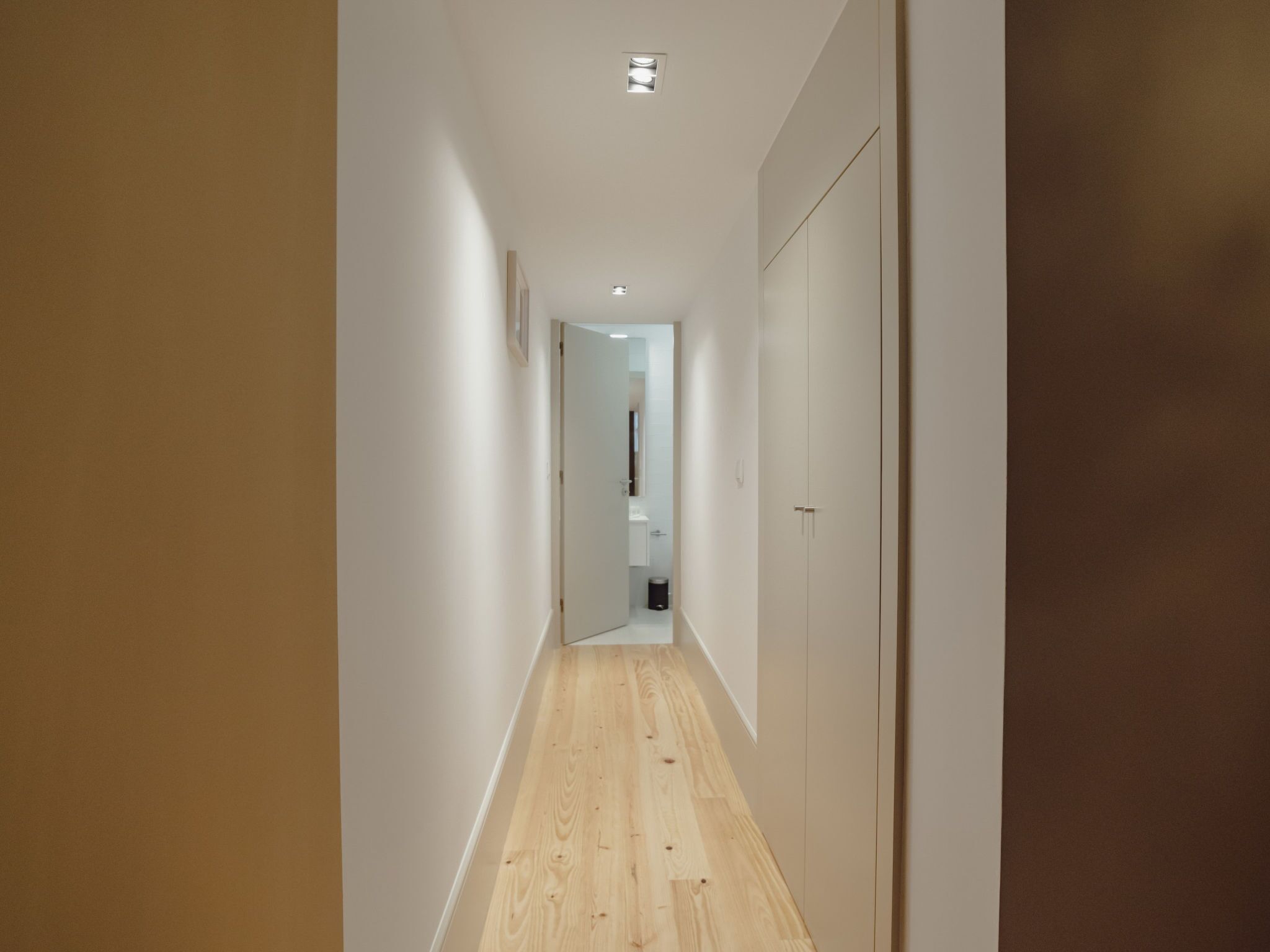 Un pasillo estrecho con piso de madera y paredes blancas, que conduce a una habitación con una puerta y un cesto de basura.