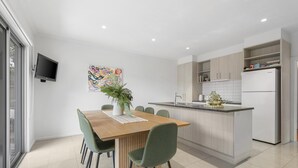 A modern kitchen with a central island, a dining area with a wooden table and green chairs, and a TV mounted on the wall. - 55 Seconds of Summer by Ready Set Stay (Rosebud)