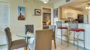 Un coin repas avec une table en verre, des chaises rembourrées, une cuisine avec des meubles blancs et un ventilateur de plafond.