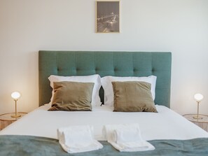 Een net opgemaakt bed met een groen gestoffeerd hoofdbord, twee bedlampen en een ingelijst schilderij aan de muur.