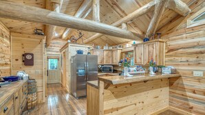 Interior de una cabaña de troncos con cocina, que incluye armarios de madera, un refrigerador, un microondas y una isla con encimera de madera.