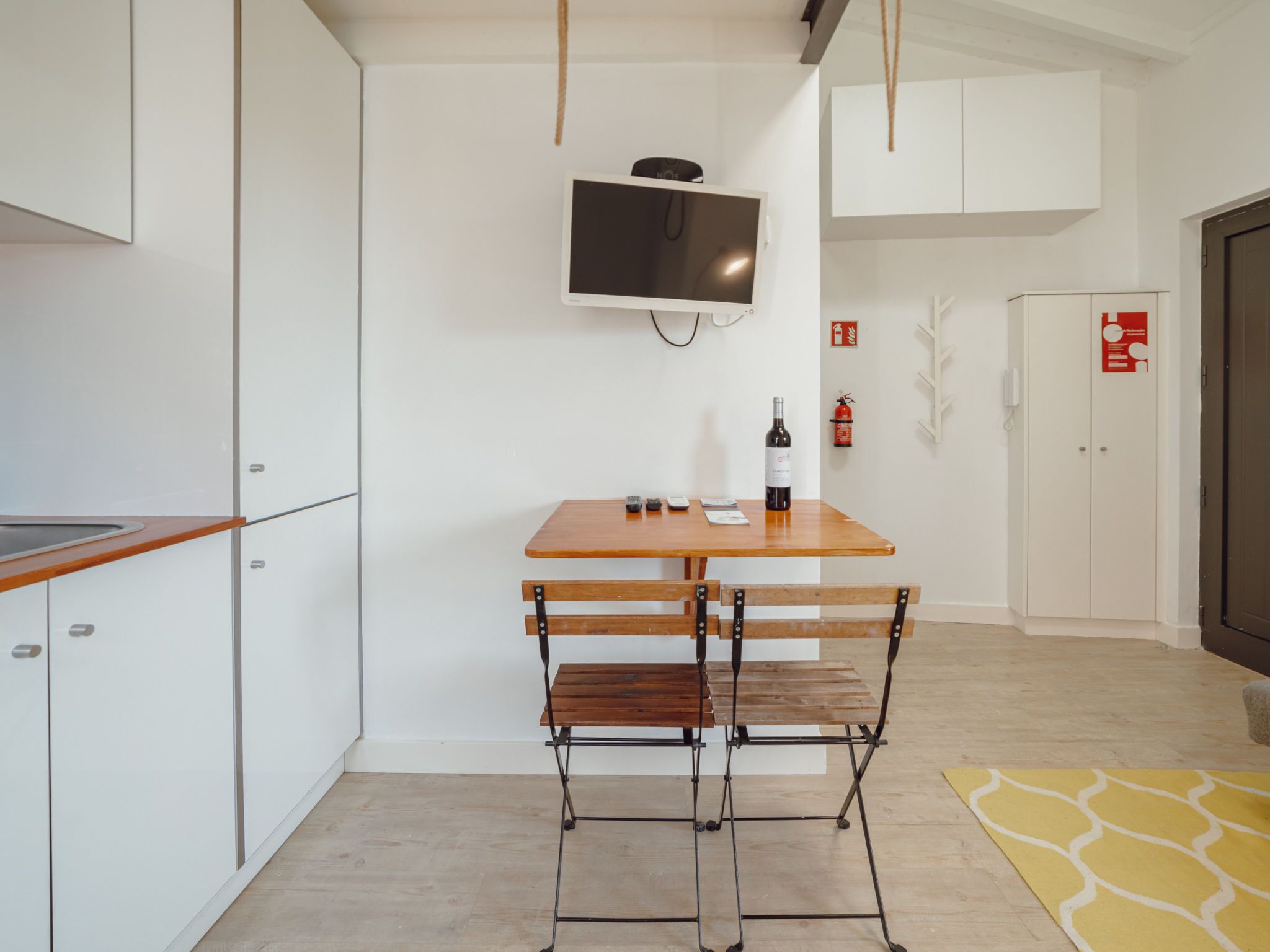 Een moderne keuken met een houten tafel, barkrukken, een wandgemonteerde televisie en een brandblusser aan de muur.