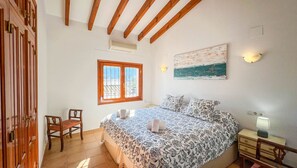 Ein Schlafzimmer mit Bett, Stuhl, Nachttisch, Lampe, einem Gemälde, einem Fenster und Holzbalken an der Decke.