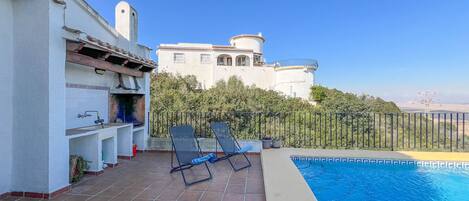 A poolside area with a tiled deck, lounge chairs, and a view of a large building with a tower.
