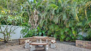 A circular outdoor seating area with a central table and benches, surrounded by a white wall and lush greenery. - Luxe Ft Lauderdale Pool Oasis Near Airport & Beach (Fort Lauderdale)
