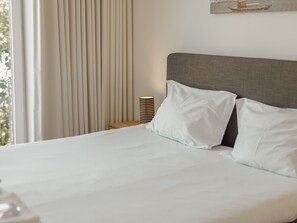 Un lit bien fait, avec du linge de lit blanc, une tête de lit grise et une lampe à rayures sur une table de chevet.