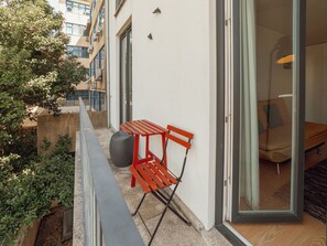 Ein Balkon mit einer roten Bank und einem großen Pflanzgefäß, mit Blick auf eine Stadtlandschaft.