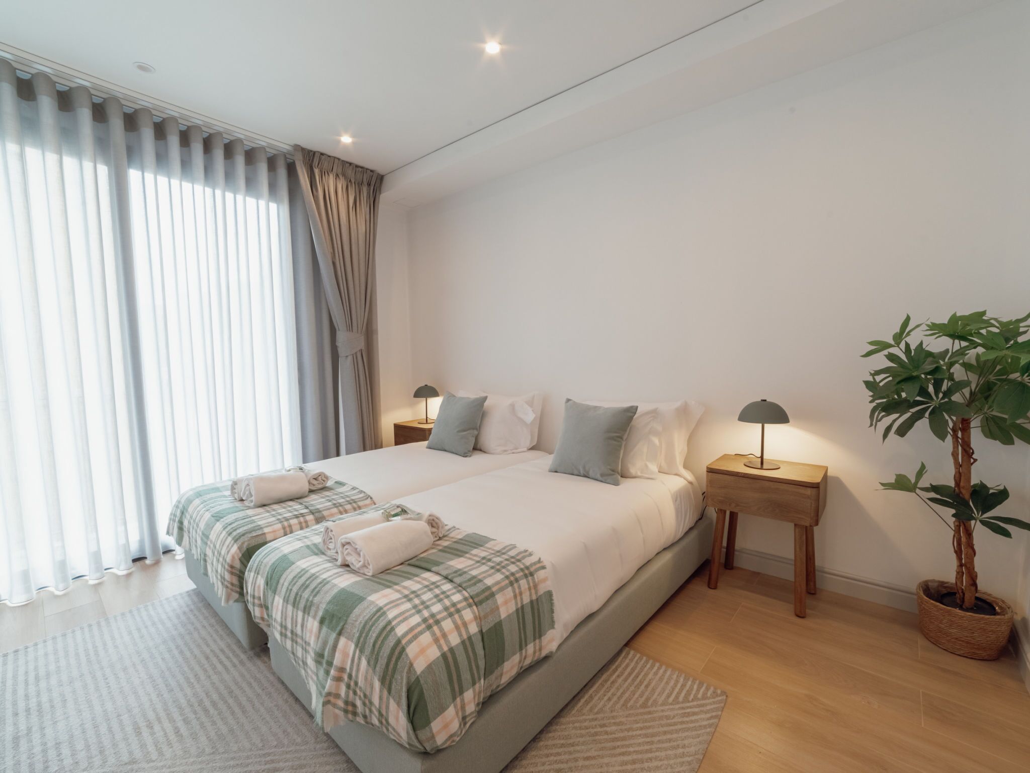 Quarto moderno com cama grande, abajures, um vaso de planta e vista para o exterior.