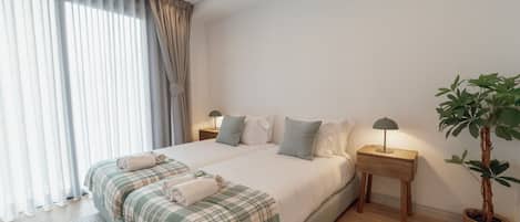 A modern bedroom with a large bed, bedside lamps, a potted plant, and a view of the outdoors.