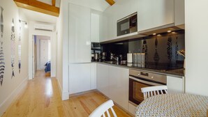 Eine moderne Küche mit weißen Schränken, einem eingebauten Backofen und einer Mikrowelle. Die Holzbalkendecke ist sichtbar, und auf dem Esstisch liegt eine Tischdecke mit Muster.