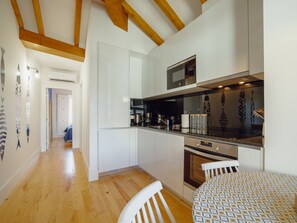 Eine moderne Küche mit weißen Schränken, einem eingebauten Backofen und einer Mikrowelle. Die Holzbalkendecke ist sichtbar, und auf dem Esstisch liegt eine Tischdecke mit Muster.