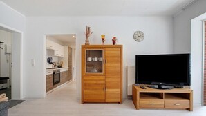 Een moderne woonkamer met een houten kast, een flatscreen televisie op een houten standaard, en een keuken op de achtergrond.
