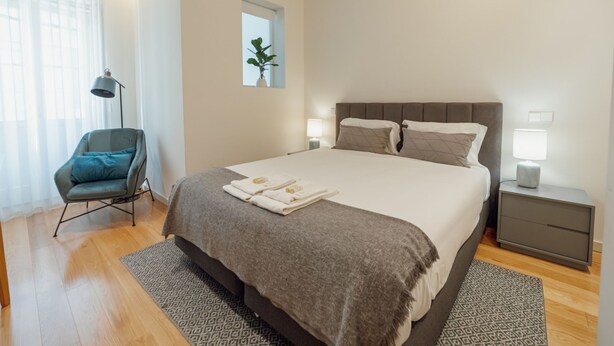 Um quarto moderno com uma cama grande, abajures, uma cadeira e uma janela pequena.