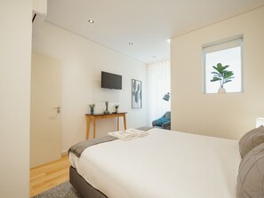 Quarto moderno com uma cama grande, uma TV de tela plana na parede, uma mesinha com objetos decorativos e uma janela com uma planta.
