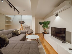 Ein modernes Wohnzimmer mit einem Ecksofa, einem kleinen runden Tisch, einem Fernseher und einer Zimmerpflanze.