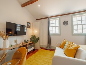 Ein modernes Wohnzimmer mit einem Flachbildfernseher, einem Sofa mit gelben Kissen, einem Holztisch und einem großen Fenster mit Vorhängen.