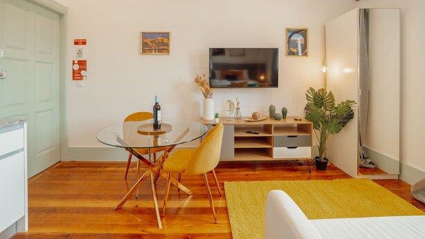Una sala moderna con una mesa de comedor de vidrio, sillas amarillas, un televisor de pantalla plana, vigas de madera, una planta grande y una alfombra amarilla.