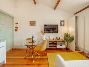 Ein modernes Wohnzimmer mit einem Glastisch, gelben Stühlen, einem Flachbildfernseher, Holzbalken, einer großen Pflanze und einem gelben Teppich.