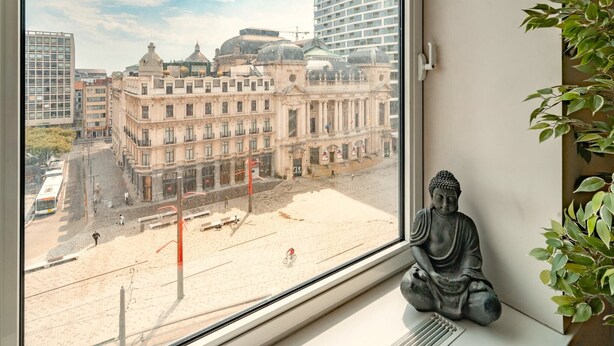 Une vue à travers la fenêtre d’un bâtiment à l’architecture classique, avec une statue de Bouddha et une plante en pot.