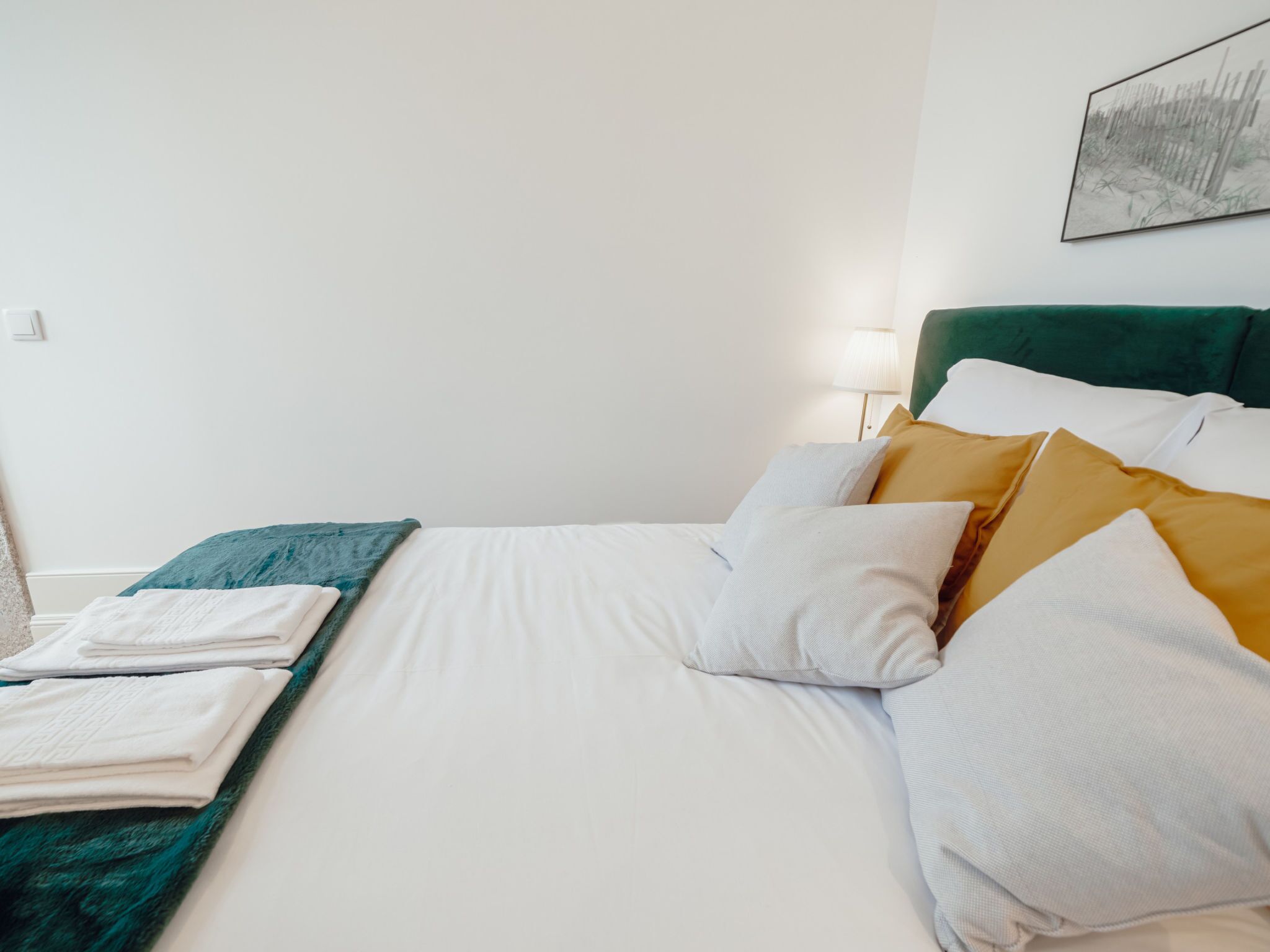 A neatly made bed with white linens and multiple pillows, a green headboard, and a lamp beside it.