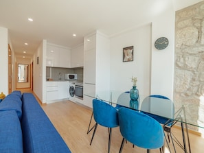 A modern kitchen with white cabinets, a microwave, and an oven. There is a dining area with a glass table and blue chairs. A clock and a leopard painting are on the wall. - Apartment  by Interhome (Porto)