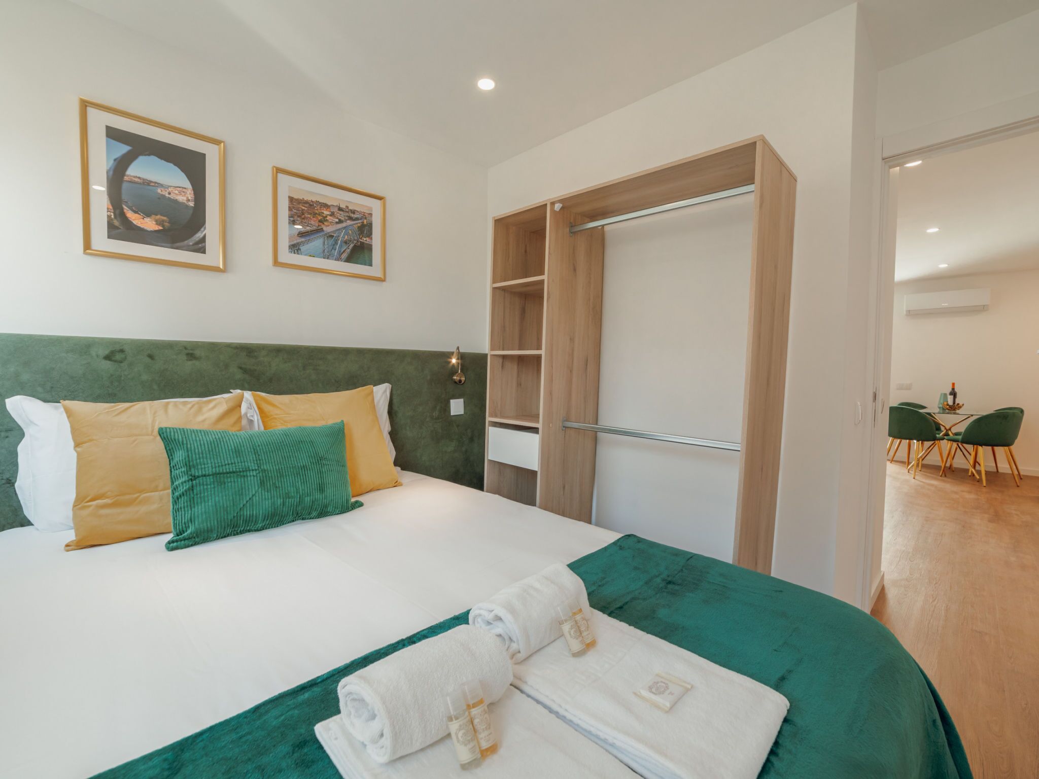 Une chambre d’hôtel avec un lit, des coussins verts et jaunes, un plaid vert, une armoire en bois et deux tableaux encadrés accrochés au mur.