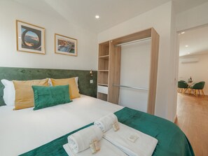 Une chambre d’hôtel avec un lit, des coussins verts et jaunes, un plaid vert, une armoire en bois et deux tableaux encadrés accrochés au mur.