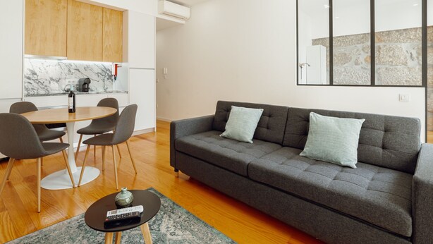 A modern living room with a grey sofa, a dining table with chairs, a coffee table with a remote control, and a kitchen area with wooden cabinets and a marble countertop. - Apartment  by Interhome (Porto)