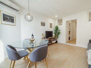 Ein modernes Wohnzimmer mit einem Glastisch, zwei grauen Stühlen, einem Flachbildfernseher, einer Topfpflanze und gerahmten Bildern an der Wand.