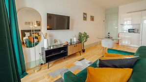 Ett modernt vardagsrum med en platt-TV, en rund spegel, en soffa med kuddar, ett soffbord och en liten köksdel med en mikrovågsugn.