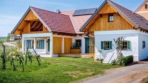 A two-story wooden house with a sloped roof, a small porch, and a garden with grapevines. - Klöchberg R&L APP Sunrise - Gelber Muskateller (Klöch)