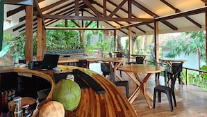 Un área de estar al aire libre con mesas y sillas de madera, una barra con una computadora portátil y vistas a zonas verdes y agua.