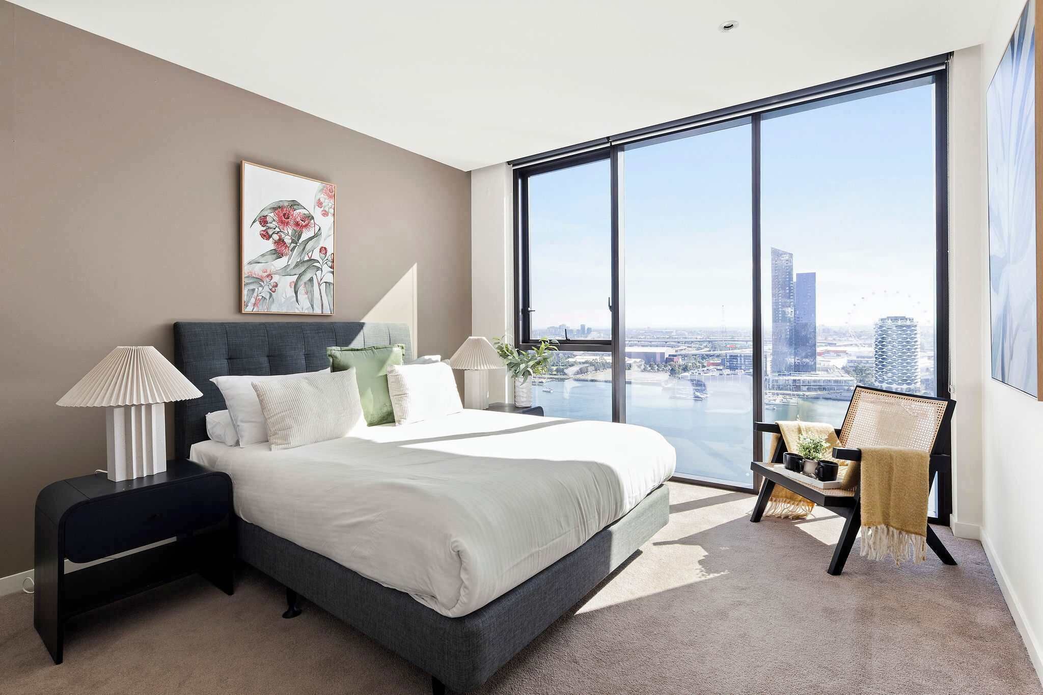 A modern bedroom with a large bed, a nightstand, a chair, and a view of a cityscape through a large window.