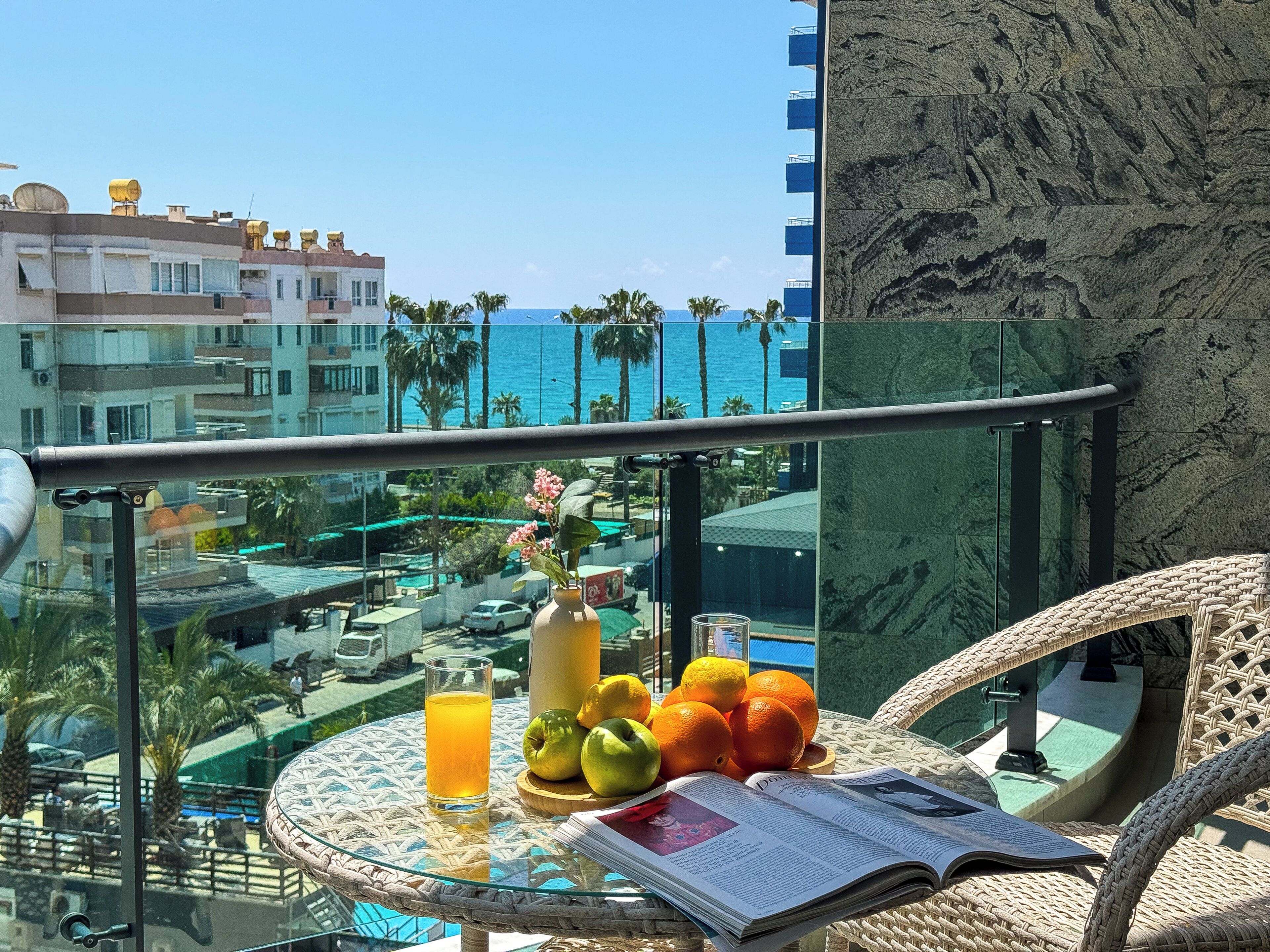 Un balcon avec une table en verre, un magazine, un verre de jus d’orange et des fruits.