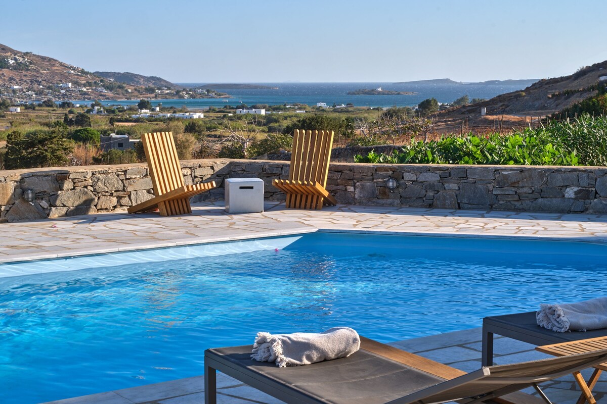 A pool with lounge chairs and a stone wall, overlooking a coastal landscape.