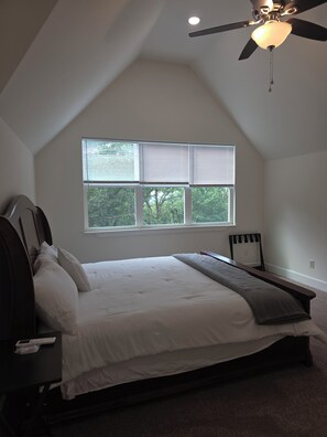 Ein Schlafzimmer mit Bett, Deckenventilator, Fenster mit Jalousien und Blick auf Bäume.