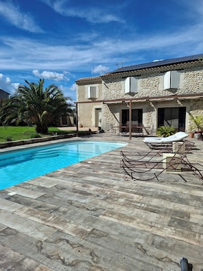 Un edificio de piedra con piscina, sillones de exterior y una palmera.