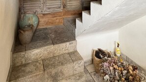 Un escalier en pierre menant à une porte en bois avec des volets, un panier tressé posé sur le palier et un petit vase contenant des fleurs séchées.