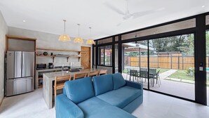 A modern kitchen with a refrigerator, stove, and dining area. There is a blue sofa in the living room and large windows with a view of the backyard. - A Secluded Modern Villa with Private Pool, Jungle Oasis Bathtub & Beach Access (puerto viejo de talamanca)