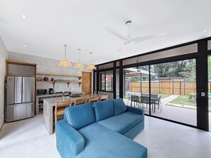 A modern kitchen with a refrigerator, stove, and dining area. There is a blue sofa in the living room and large windows with a view of the backyard. - A Secluded Modern Villa with Private Pool, Jungle Oasis Bathtub & Beach Access (puerto viejo de talamanca)